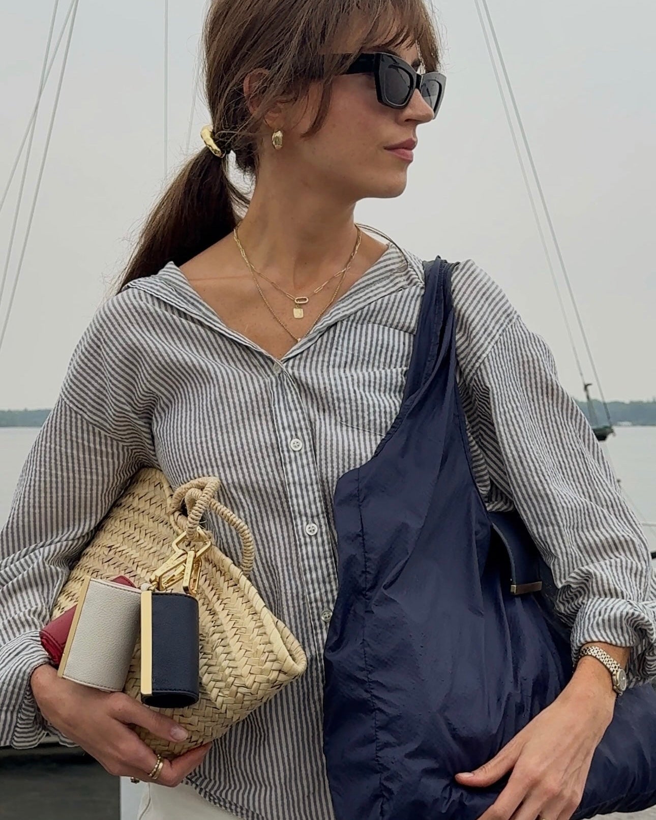 Woman holding a navy blue bag and a small woven bag, wearing sunglasses and a striped shirt.